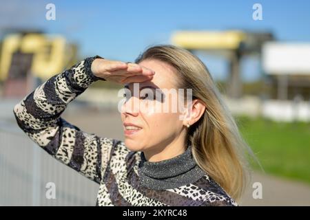 Eine attraktive blonde Frau mittleren Alters, die in der Sonne steht und ihre Hand über den Augen hält, um in die Ferne zu schauen, gegen eine Schleuse Stockfoto