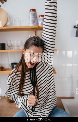 Lustige Frau, die in den Teigschaber singt und den Teigschaber als Mikrofon hält. Stockfoto