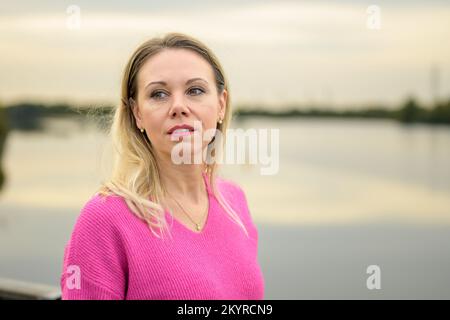 Eine wunderschöne Frau mittleren Alters mit pinkfarbenem Pullover und pinkfarbenem Lippenstift steht im Herbst vor einem Fluss oder See Stockfoto