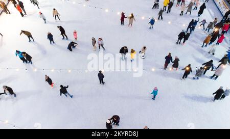 Luftaufnahme viele Leute, die im Winter auf einer Freiluft-Eisbahn Schlittschuhlaufen. Eislaufen, Draufsicht. Eisbahn Im Stadtpark. Outdoor-Aktivitäten im Winter. Hintergrundinformationen zu Eislaufferien. Weihnachtsbeleuchtung am Neujahrstag Stockfoto