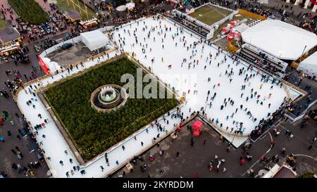 Viele Leute, die im Winter auf einer weißen Eislaufbahn im Freien Schlittschuhlaufen. Draufsicht. Hintergrund der Weihnachtsfeiertage im Neujahr. Panoramablick auf die Stadt. Lifestyle-Freizeit, aktiver Spaß Stockfoto