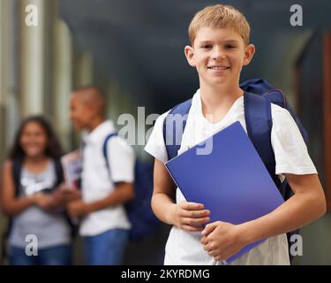 Er ist ein fleißiger Schüler. Porträt eines glücklichen Schuljungen, der im Korridor steht, mit seinen Freunden im Hintergrund. Stockfoto