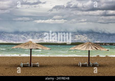 Nin, Kroatien - zwei Schilf-Sonnenschirme am leeren Queen's Beach in der mediterranen Stadt Nin am Ende des Sommers mit Velebit-Bergen im Hintergrund Stockfoto