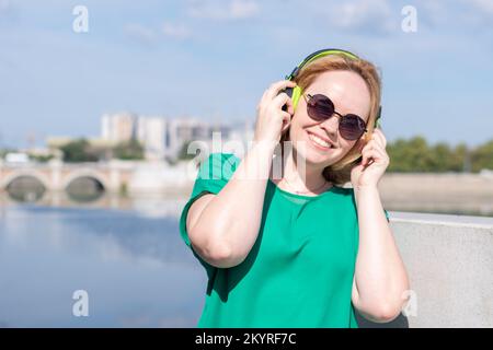 Porträt eines lächelnden, glücklichen Mädchens in Sonnenbrillen und Kopfhörern, das am Flussufer Musik hört. Ein Mädchen gegen den Himmel, eine Kopie des Raumes. Ein Mädchen wa Stockfoto
