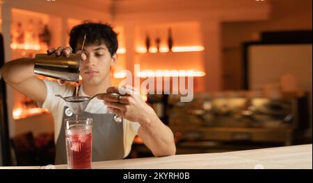 Der Mann Barkeeper filtert den hellen alkoholischen Cocktail aus einem Stahlschüttler durch ein Sieb in das Glas. Nahaufnahme. Stockfoto