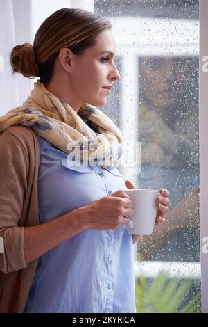 An einem Wintertag warm zu bleiben. Eine junge Frau, die aus ihrem Fenster schaute, während sie eine Tasse Kaffee hielt. Stockfoto