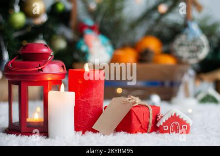 Weihnachtskerzen und Geschenk mit Anhängern zum Schreiben. Im Hintergrund befinden sich ein Weihnachtsbaum und Geschenke mit Lichtern. Neujahrshintergrund. Stockfoto