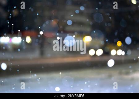 Unscharfer Hintergrund. Blick auf die Stadt, Lichter, fallender Schnee, Nacht, Straße, Bokeh die Flecken der Scheinwerfer der sich bewegenden Autos. Diffuse städtische Kulisse Winterlandschaft der Straße in der Stadt bei Nacht. Laterne Licht, Schneefall Stockfoto