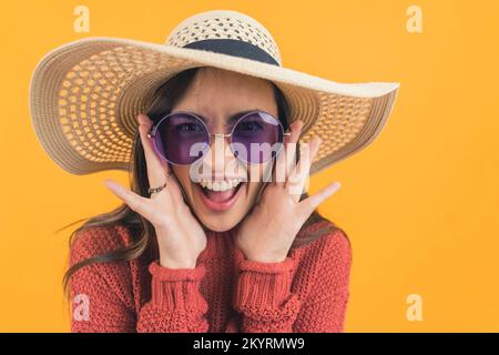 Aufgeregte junge Erwachsene lateinamerikanische Frau, die eine Sonnenbrille und einen Strohhut trägt und Hände an Gesicht hält und mit einem breiten Lächeln in die Kamera schaut. Sommerferien. Horizontales Studiofoto mit gelbem Hintergrund. Hochwertiges Foto Stockfoto
