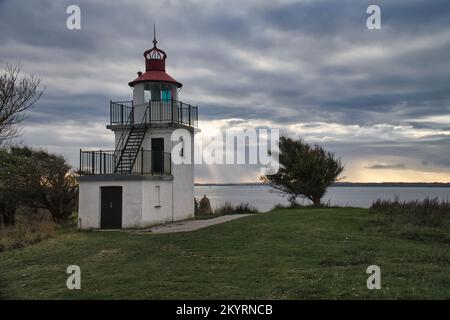 Leuchtturm, Spodsbjerg Fyr in Huntsted an der Küste Dänemarks. Die Sonne scheint durch die Wolken. Wiese mit Bäumen. Landschaftsfoto vom Meer Stockfoto