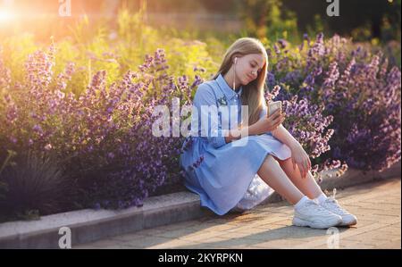 Die wunderschöne blonde Frau hört Musik über Kopfhörer auf ihrem Smartphone und sitzt in Blumen Stockfoto