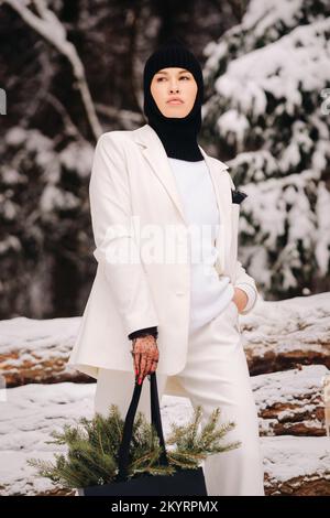 Ein Mädchen in einem weißen Anzug und Balaklava mit einem Paket Weihnachtsbäume im Winterwald an Silvester. Neujahrskonzept Stockfoto