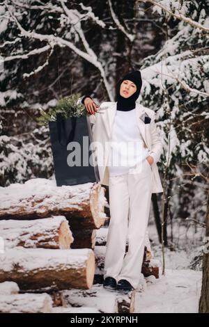 Ein Mädchen in einem weißen Anzug und Balaklava mit einem Paket Weihnachtsbäume im Winterwald an Silvester. Neujahrskonzept Stockfoto