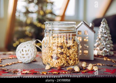 Konzept eines hausgemachten Weihnachtsgeschenks, handgemachtes geröstetes Müsli mit verschiedenen Nüssen und Rosinen in einem wunderschönen Glasgefäß. Verwischen Sie Weihnachtsbaum und Lichter. Stockfoto