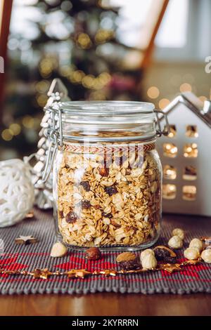 Konzept eines hausgemachten Weihnachtsgeschenks, handgemachtes geröstetes Müsli mit verschiedenen Nüssen und Rosinen in einem wunderschönen Glasgefäß. Verwischen Sie Weihnachtsbaum und Lichter. Stockfoto