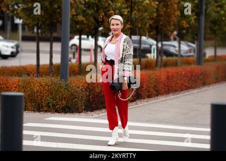 Modetrends für den Herbst. Frau mit kurzer Frisur, die über die Straße läuft, rote Hosen, gestreiftes Hemd, Strickjacke, weiße Schuhe und schwarz trägt Stockfoto