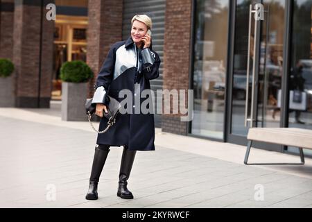 Modefrau in trendigen Outfits, schwarzer Mantel, Lederstiefel, Leggings. Das stilvolle, modische Modell spricht und lächelt auf der Straße in der Nähe Stockfoto