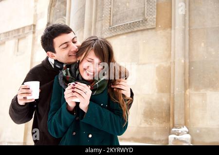Trinken Sie bei kaltem Winterwetter auf der Straße heißen Kräutertee in Pappbechern für zwei Personen. Umarmen, küssen, Spaß haben und töricht sein Stockfoto