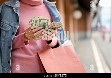 Wunderschöne und reiche Frau, die US-Dollar-Banknoten in der Hand hält, ihr Geld zählt, während sie im Einkaufszentrum einkauft. Zugeschnittenes Bild Stockfoto