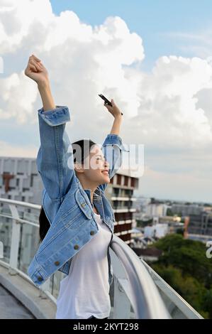 Porträt, wunderschöne junge asiatische Frau, Hände hoch in die Luft, hebt die Hand, atmet tief durch die frische Luft, steht auf dem Dach des Gebäudes. Stockfoto