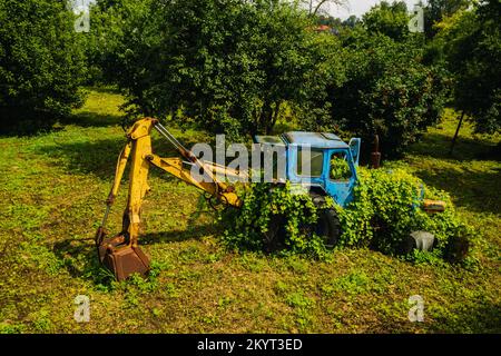 Ein alter Traktor mit Eimer, verlassen von einem grünen Garten Stockfoto