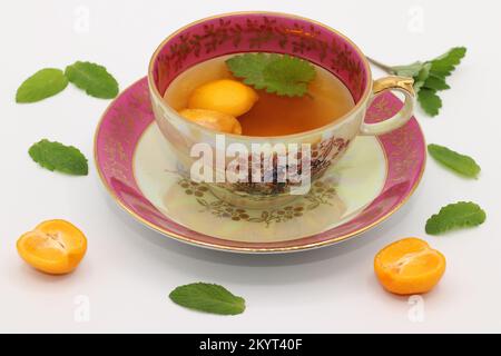 Eine wunderschöne klassische Porzellan-Teetasse mit melissa-Blatt und Calamansi-Scheiben. Minzeblätter und Calamondin-Fruchthälften liegen herum. Citrus microcarpa Stockfoto