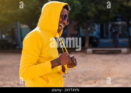 Lächelnder junger afrikanischer Mann mit Sonnenbrille und gelbem Sweatshirt mit Kapuze, die er auf dem Kopf trägt. Er ist auf der Straße und greift Stockfoto