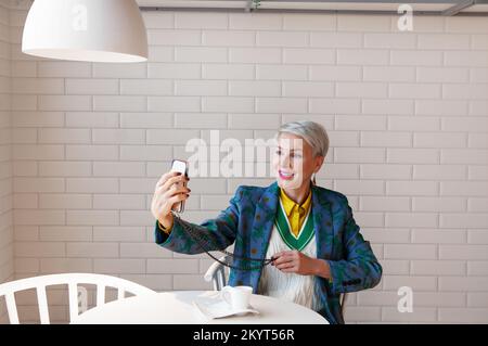 Stilvolle ältere Frau schaut auf den Smartphone-Bildschirm und lächelt für Online-Selfie-Chats. Business-Frau hält Handy in der Tasche mit Kettenband. Mode Stockfoto