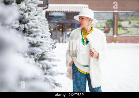 Winter helle Mode im Street-Stil einer Frau mittleren Alters, die eine Fedora-Mütze und einen weißen Kunstpelzmantel mit bunten Details trägt Stockfoto