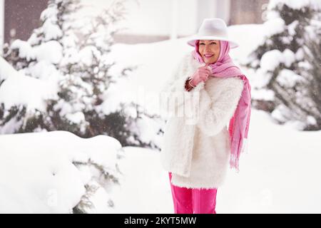 Lächelnde Erwachsene Frau vor dem Hintergrund des Schnees in modischer warmer Kleidung, Winterjacke, Schal auf dem Kopf, Fedora-Hut Stockfoto