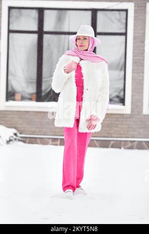 Modische, reife Frau in pinkfarbenem Business-Anzug mit Hose, Kopftuch, weißer Filzmütze, Kunstpelzmantel und Winter-Outfit im Street-Stil Stockfoto