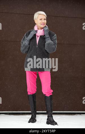 Winteroutfit für ältere Frauen mit hohen Stiefeln, pinkfarbenen Hosen, Bomberjacke, stilvollem, durchgehendem Look einer älteren Frau mit kurzen Haaren und leuchtenden Details Stockfoto