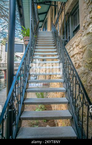 Treppe, die ein Gebäude entlang des San Antonio River Walk in Texas hinaufführt. Die malerische Landschaft des Flusses, der Brücke und der Gebäude ist in t zu sehen Stockfoto