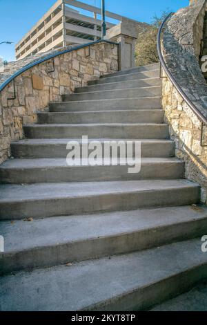 Außentreppe mit Steinhalmwand und Geländern am San Antonio River Walk. Blick vom Boden einer Parktreppe mit Gebäuden vor blauem Himmel Stockfoto