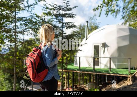 Frau, die sich die Geo-Dome-Zelte der Natur ansieht. Grüner, blauer, orangefarbener Hintergrund. Gemütlich, Camping, Glamping, Urlaub, Urlaub Lifestyle-Konzept. Landschaftlich reizvolle Natur Stockfoto