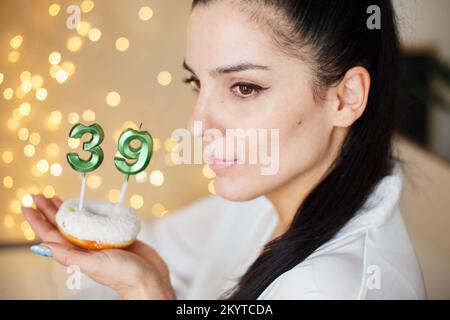 Eine Frau, die einen Kuchen mit den Kerzen Nummer 39 auf festlichem, verschwommenem Bokeh-Hintergrund hält. Stockfoto
