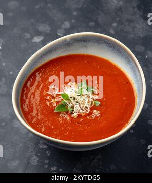 Schöne Keramikschüssel voller cremiger und würziger Tomatensuppe, garniert mit geriebenem Käse und Basilikum Stockfoto