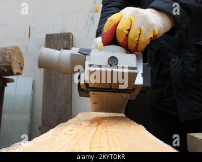 Die Arbeit mit einer elektrischen Hobelmaschine auf einer soliden Holzplatte glättet und betont die natürliche Textur von Holz, eine Hobelmaschine in den Handschuhen des Zimmermanns Stockfoto
