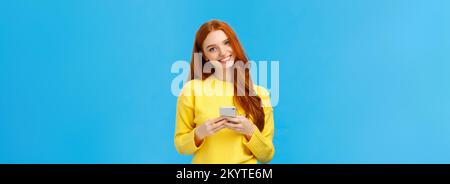 Technologie-, Internet- und Gadgets-Konzept. Süße Rotkopffrau, die Textfreundin, Messaging, Unterhaltung mit Smartphone-App, lächelnde Kamera schickt Stockfoto