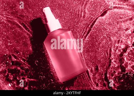 Matte Glasflasche mit Körperöl auf Viva Magenta Farbe Wasserhintergrund, Glaskosmetik im Wasser, Draufsicht. Farbe des Jahres 2023 Stockfoto