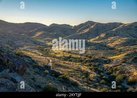 Phoenix, Arizona - Pima Canyon Trail in einem Tal vor der Skyline des Sonnenuntergangs. Zwischen den Bergketten gibt es Wanderwege mit einem grünen Wildplan Stockfoto