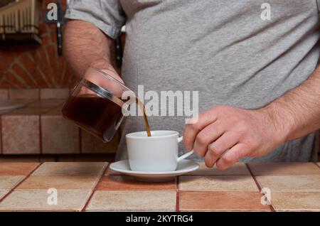 Männliche Hände gießen Kaffee in eine Kaffeetasse, die auf einem gefliesten Küchentisch steht Stockfoto