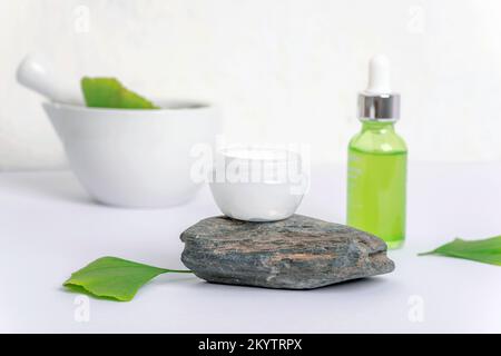 Frische grüne Ginkgoblätter, Mörtel und Stößel, kosmetische Serumflasche und Cremebecher auf weißem Hintergrund. Natürliches, handgefertigtes Kosmetikkonzept. Selektiv Stockfoto