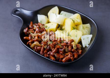 Pfifferling-Pilze mit gekochten Kartoffeln, Zwiebeln, Dill und Öl in einer kleinen schwarzen Pfanne. Traditionelles russisches Gericht. Schwarzer Hintergrund, Nahaufnahme. Stockfoto