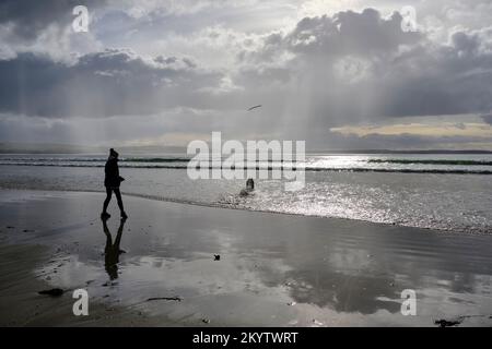 Eine Frau an einem beleuchteten Strand in Dunnet Bay, die einen Stock für einen Hund wirft, der im Meer wartet, um ihn zu holen Stockfoto