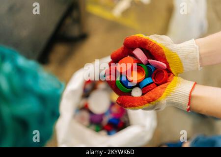 Nahaufnahme aus dem Hochwinkel von unbekannten weiblichen Freiwilligen in Handschuhen mit einer Handvoll mehrfarbiger Kunststoffdeckel zur weiteren Entsorgung. Stockfoto