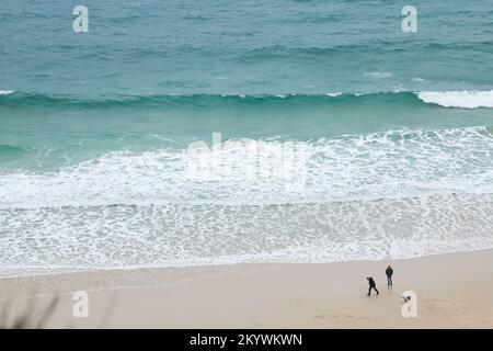 Ein Paar mit seinem Hund am Strand in Porthcurno Bay, Cornwall, England, neben dem türkisfarbenen Wasser des Ozeans. Stockfoto