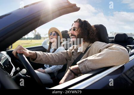 Ein junges, fröhliches weißes Paar, das in einem Cabrio fährt und die Freiheit des Wochenendausflugs genießt Stockfoto