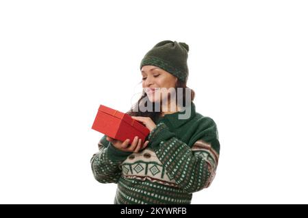 Die hübsche afroamerikanische Frau in gemustertem Xmas Pullover und grünem Hut hält ein Weihnachtsgeschenk vor weißem Hintergrund Stockfoto