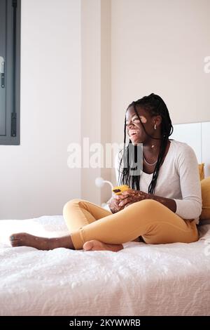 Fröhliche schwarze Frau mit Vitiligo, die auf dem Bett sitzt und ein Handy benutzt Stockfoto